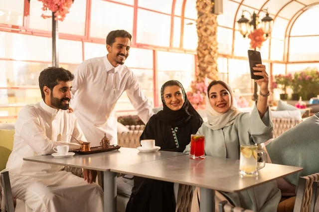 استخدام اتكنولوجيا والتقنيات الحديثة، توثيق اللحظات السعيدة من خلال التقاط الصور التذكارية، إمرأة عربية خليجية سعودية ترتدي العباءة والحجاب تجلس على الأريكة مع أصدقائها بإحدى المقاهي وتستخدم الهاتف، مشاركة المشروبات الباردة والحلويات