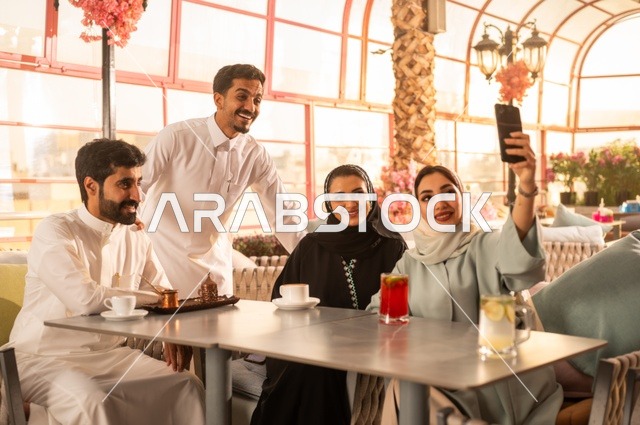 استخدام اتكنولوجيا والتقنيات الحديثة، توثيق اللحظات السعيدة من خلال التقاط الصور التذكارية، إمرأة عربية خليجية سعودية ترتدي العباءة والحجاب تجلس على الأريكة مع أصدقائها بإحدى المقاهي وتستخدم الهاتف، مشاركة المشروبات الباردة والحلويات