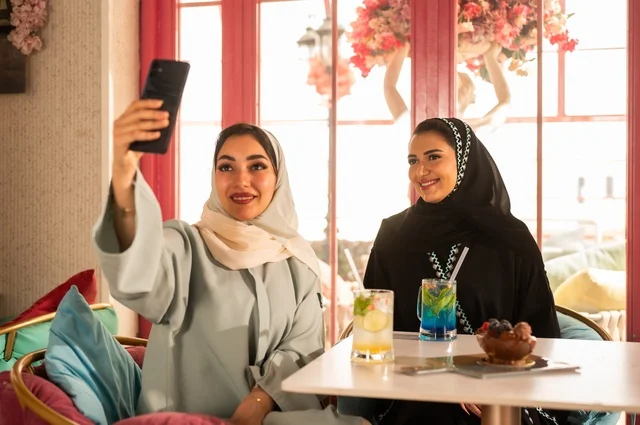 توثيق اللحظات السعيدة من خلال التقاط الصور التذكارية، إمرأة عربية خليجية سعودية ترتدي العباءة والحجاب تجلس على الأريكة مع صديقتها بإحدى المقاهي وتستخدم الهاتف، مشاركة المشروبات اللذيذة والحلويات، استخدام التكنولوجيا والتقنيات الحديثة