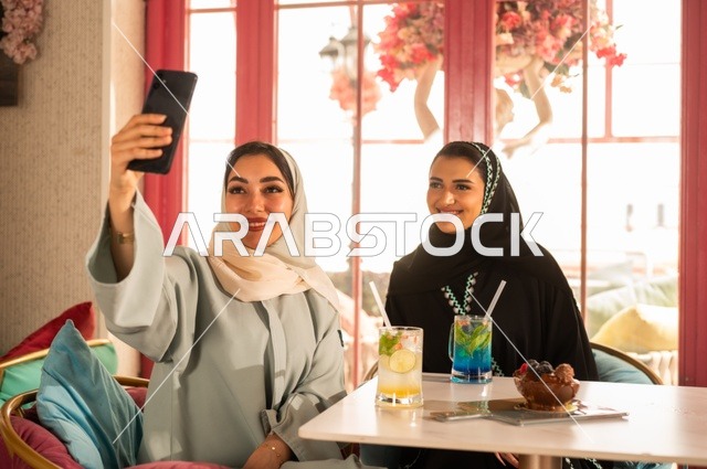 توثيق اللحظات السعيدة من خلال التقاط الصور التذكارية، إمرأة عربية خليجية سعودية ترتدي العباءة والحجاب تجلس على الأريكة مع صديقتها بإحدى المقاهي وتستخدم الهاتف، مشاركة المشروبات اللذيذة والحلويات، استخدام التكنولوجيا والتقنيات الحديثة