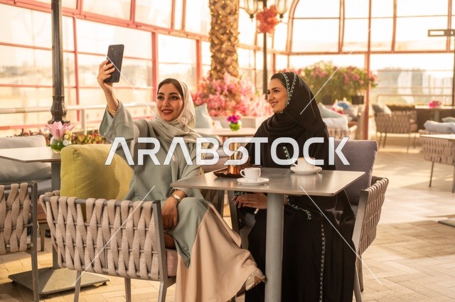توثيق اللحظات السعيدة من خلال التقاط الصور التذكارية، إمرأة عربية خليجية سعودية ترتدي العباءة والحجاب تجلس على الأريكة مع صديقتها بإحدى المقاهي وتستخدم الهاتف، مشاركة المشروبات اللذيذة والحلويات، استخدام التكنولوجيا والتقنيات الحديثة
