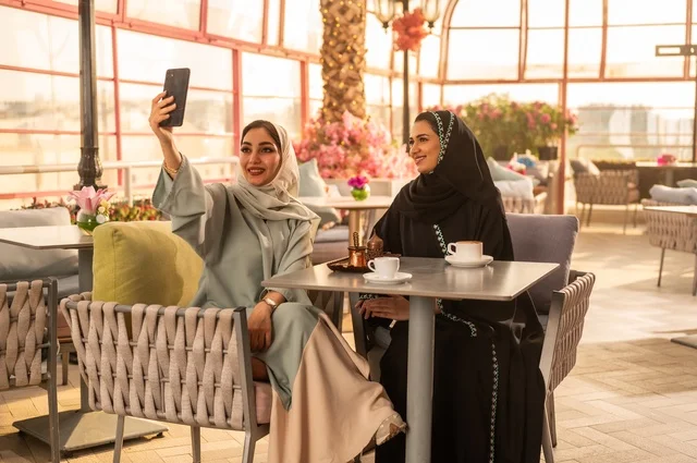 توثيق اللحظات السعيدة من خلال التقاط الصور التذكارية، إمرأة عربية خليجية سعودية ترتدي العباءة والحجاب تجلس على الأريكة مع صديقتها بإحدى المقاهي وتستخدم الهاتف، مشاركة المشروبات اللذيذة والحلويات، استخدام التكنولوجيا والتقنيات الحديثة