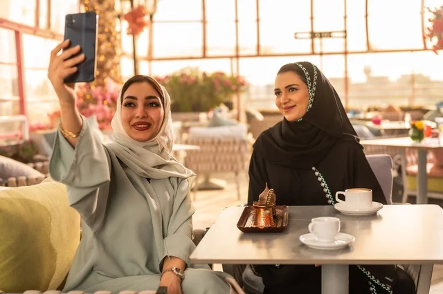Documenting happy moments through taking commemorative photos, a Gulf Arab Saudi woman wearing an abaya and hijab sits on a sofa with her friend in a café and uses her phone, sharing delicious drinks and sweets, utilizing modern technology and techniques.