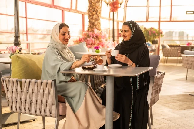 Enjoying Arabic coffee, discussing and conversing in a calm atmosphere, building successful social relationships, a Saudi Arabian Gulf woman in a black abaya sitting with her friend in a café, two Saudi young women spending enjoyable and entertaining times.