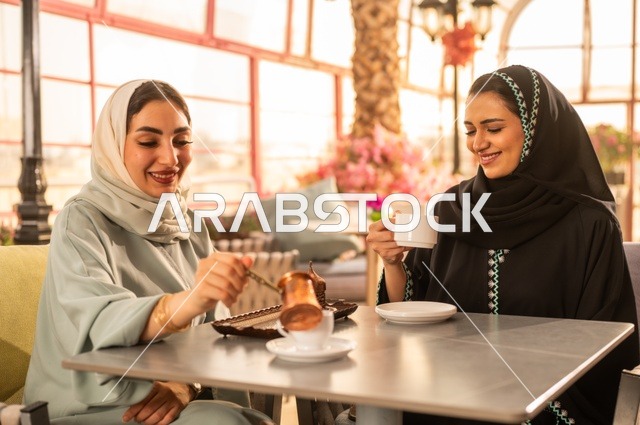 الاستمتاع بشرب القهوة العربية, النقاش والحوار في الأجواء الهادئة، بناء علاقات اجتماعية ناجحة، امرأة عربية خليجية سعودية محجبة ترتدي العباءة السوداء تجلس مع صديقتها بإحدى المقاهي, شابتان سعوديتان تقضيان أوقات ممتعة ومسلية