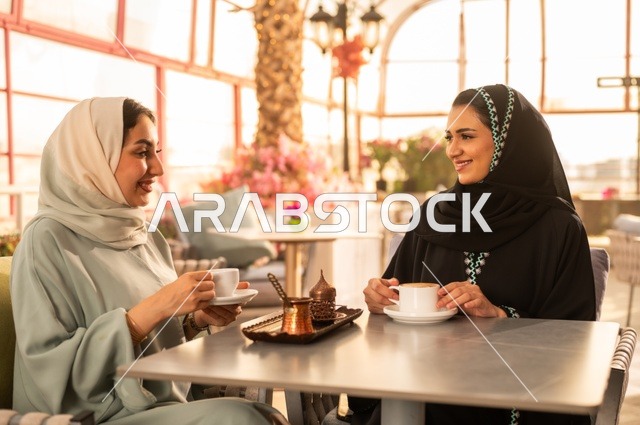 الاستمتاع بشرب القهوة العربية, النقاش والحوار في الأجواء الهادئة، بناء علاقات اجتماعية ناجحة، امرأة عربية خليجية سعودية محجبة ترتدي العباءة السوداء تجلس مع صديقتها بإحدى المقاهي, شابتان سعوديتان تقضيان أوقات ممتعة ومسلية