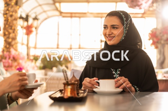 الاستمتاع بشرب القهوة العربية, النقاش والحوار في الأجواء الهادئة، بناء علاقات اجتماعية ناجحة، امرأة عربية خليجية سعودية محجبة ترتدي العباءة السوداء تجلس مع صديقتها بإحدى المقاهي, شابتان سعوديتان تقضيان أوقات ممتعة ومسلية