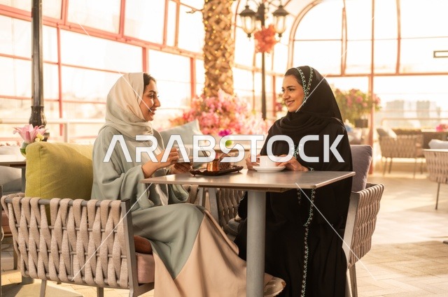 الاستمتاع بشرب القهوة العربية, النقاش والحوار في الأجواء الهادئة، بناء علاقات اجتماعية ناجحة، امرأة عربية خليجية سعودية محجبة ترتدي العباءة السوداء تجلس مع صديقتها بإحدى المقاهي, شابتان سعوديتان تقضيان أوقات ممتعة ومسلية