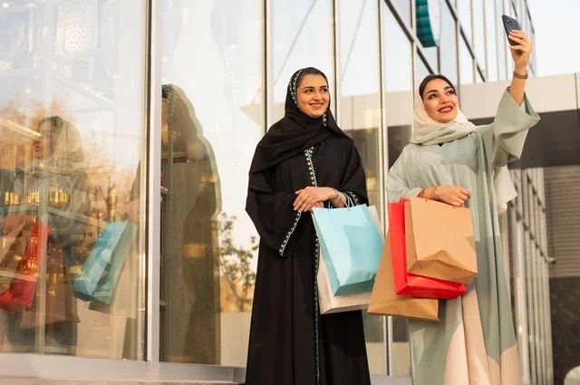 Taking selfies using a mobile phone, shopping during sales and discounts, enjoying shopping at the mall, the fun of shopping on Black Friday with friends, two Arab Gulf Saudi women wearing abayas and hijabs carrying shopping bags with expressions and gestures of happiness and joy.