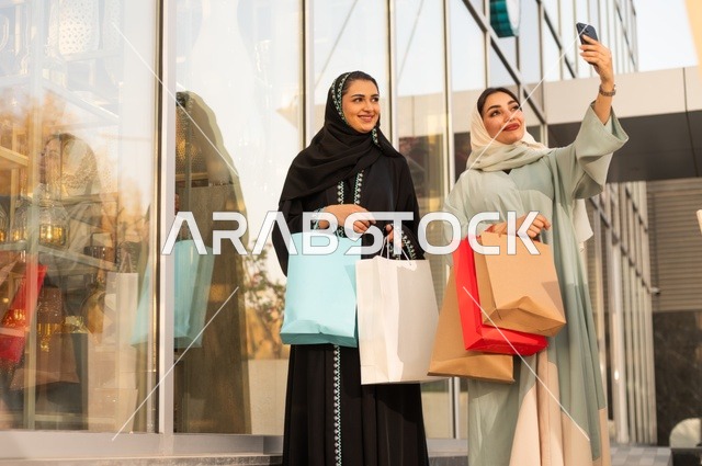 Taking selfies using a mobile phone, shopping during sales and discounts, enjoying shopping at the mall, the fun of shopping on Black Friday with friends, two Arab Gulf Saudi women wearing abayas and hijabs carrying shopping bags with expressions and gestures of happiness and joy.