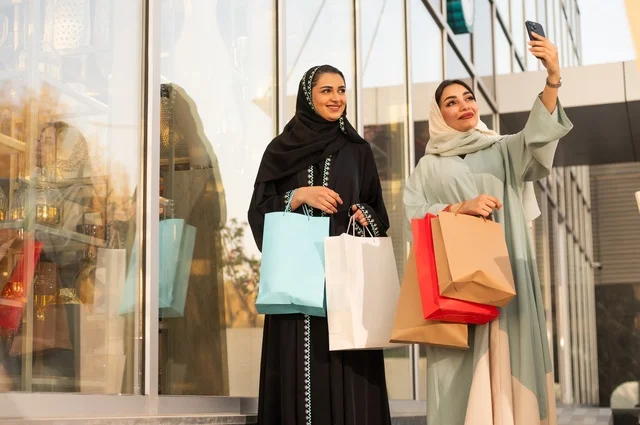 Taking selfies using a mobile phone, shopping during sales and discounts, enjoying shopping at the mall, the fun of shopping on Black Friday with friends, two Arab Gulf Saudi women wearing abayas and hijabs carrying shopping bags with expressions and gestures of happiness and joy.