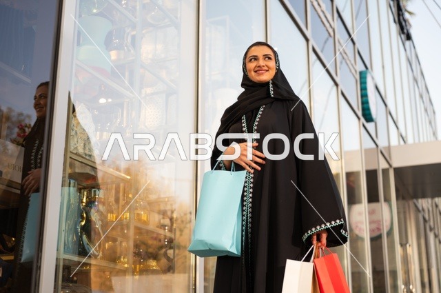 استغلال العروض والتخفيضات الموسمية والجمعة البيضاء، التسوق من المراكز التجارية  ، اختيار المستلزمات من المول التجاري، سيده عربية خليجية سعودية ترتدي العباءه و الحجاب تحمل بيدها أكياس ورقية