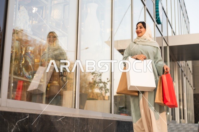 Shopping from shopping centers, selecting supplies from the mall, a Saudi Gulf Arab woman wearing an abaya and hijab carrying paper bags in her hand, taking advantage of seasonal offers and discounts and Black Friday.