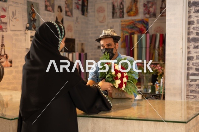 Shops and stores selling flowers and natural flowers for holidays and occasions, a Saudi Arabian Gulf woman wearing an abaya and hijab buying a bouquet of natural red roses from the seller, gestures indicating joy and happiness