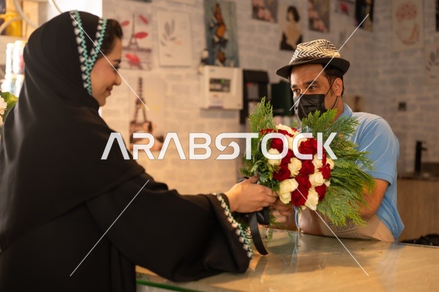 متاجر ومحلات بيع الورود والزهور الطبيعية للأعياد والمناسبات, امرأة عربية خليجية سعودية ترتدي العباءة والحجاب تقوم بشراء باقة ورد احمر طبيعي من البائع , ايماءات تدل على الفرح والسعاده