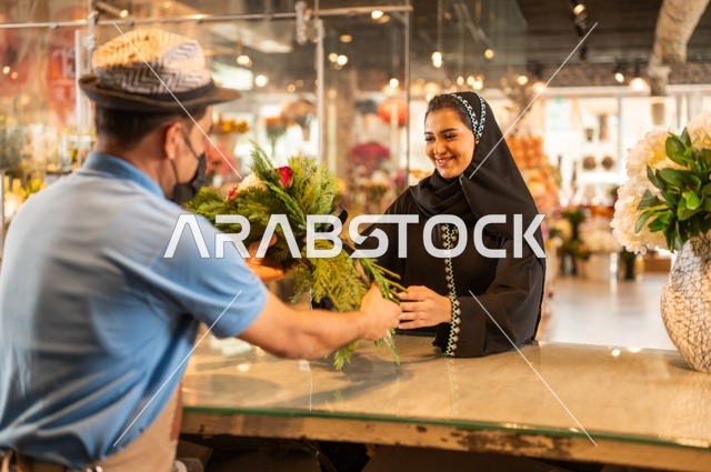 متاجر ومحلات بيع الورود والزهور الطبيعية للأعياد والمناسبات, امرأة عربية خليجية سعودية ترتدي العباءة والحجاب تقوم بشراء باقة ورد احمر طبيعي من البائع , ايماءات تدل على الفرح والسعاده