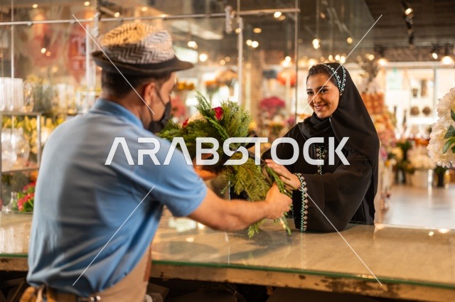 متاجر ومحلات بيع الورود والزهور الطبيعية للأعياد والمناسبات, امرأة عربية خليجية سعودية ترتدي العباءة والحجاب تقوم بشراء باقة ورد احمر طبيعي من البائع , ايماءات تدل على الفرح والسعاده