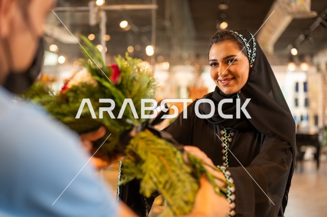 متاجر ومحلات بيع الورود والزهور الطبيعية للأعياد والمناسبات, امرأة عربية خليجية سعودية ترتدي العباءة والحجاب تقوم بشراء باقة ورد احمر طبيعي من البائع , ايماءات تدل على الفرح والسعاده