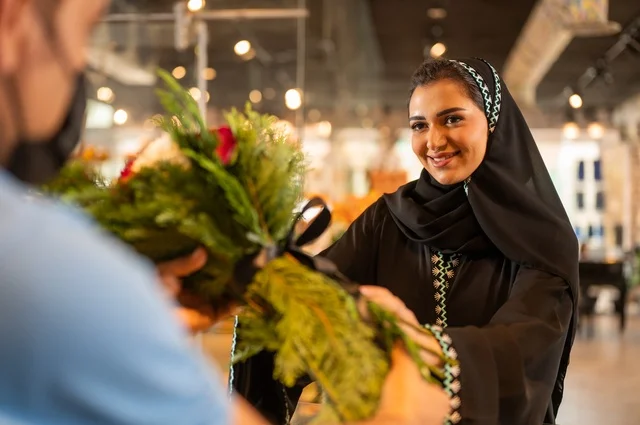 متاجر ومحلات بيع الورود والزهور الطبيعية للأعياد والمناسبات, امرأة عربية خليجية سعودية ترتدي العباءة والحجاب تقوم بشراء باقة ورد احمر طبيعي من البائع , ايماءات تدل على الفرح والسعاده