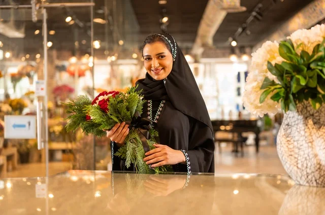 محلات ومتاجر بيع وشراء الورود والزهور الطبيعية للأعياد والمناسبات, امرأة عربية خليجية سعودية ترتدي العباءة والحجاب تحمل باقة ورد احمر طبيعي تنظر الى الكاميرا بايماءات تدل على الفرح والسعاده