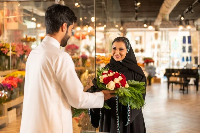 Expressions and gestures of affection and happiness, shops and stores for buying and selling natural flowers and roses for holidays and occasions, a Saudi Arabian Gulf husband wearing traditional attire presents a bouquet of natural red roses to his wife.