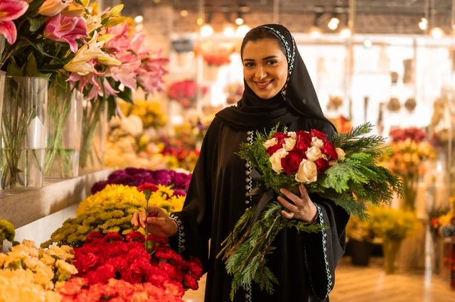 شراء الورود والزهور الطبيعية للأعياد والمناسبات من المحلات والمتاجر, امرأة عربية خليجية سعودية ترتدي العباءة والحجاب تحمل باقة ورد احمر طبيعي بايماءات الفرح والسعاده