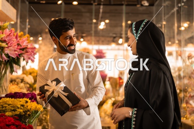 زوج عربي خليجي سعودي يرتدي الثوب التقليدي يقوم بإهداء باقة ورد احمر طبيعي وهدية مغلفة لزوجته، تعابير وايماءات المودة والسعاده، محلات ومتاجر بيع وشراء الورود والزهور الطبيعية للأعياد والمناسبات
