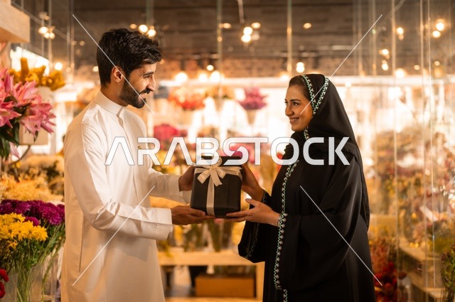 زوج عربي خليجي سعودي يرتدي الثوب التقليدي يقوم بإهداء باقة ورد احمر طبيعي وهدية مغلفة لزوجته، تعابير وايماءات المودة والسعاده، محلات ومتاجر بيع وشراء الورود والزهور الطبيعية للأعياد والمناسبات