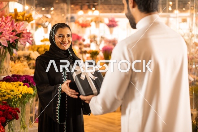 زوج عربي خليجي سعودي يرتدي الثوب التقليدي يقوم بإهداء باقة ورد احمر طبيعي وهدية مغلفة لزوجته، تعابير وايماءات المودة والسعاده، محلات ومتاجر بيع وشراء الورود والزهور الطبيعية للأعياد والمناسبات