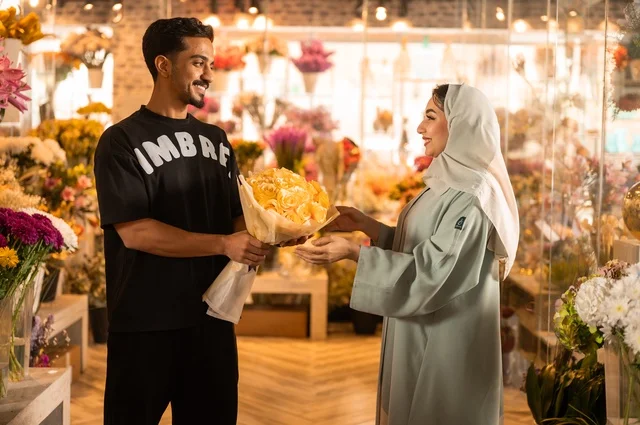Celebrating holidays and special occasions, a Saudi Gulf Arab husband dressed in casual attire presents a bouquet of natural yellow flowers to his wife, expressions and gestures of affection and happiness, shops and stores for buying and selling flowers and natural blooms.