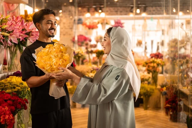 Celebrating holidays and special occasions, a Saudi Gulf Arab husband dressed in casual attire presents a bouquet of natural yellow flowers to his wife, expressions and gestures of affection and happiness, shops and stores for buying and selling flowers and natural blooms.