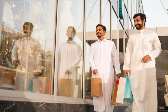 المتعة في التسوق واقتناء المستلزمات والهدايا، الاستفادة من العروضات والتخفيضات، رجلان عربيان خليجيان سعوديان يرتديان الشماغ والثوب التقليدي يحملان بيده أكياس ورقية ملونة بإيماءات السرور والسعادة