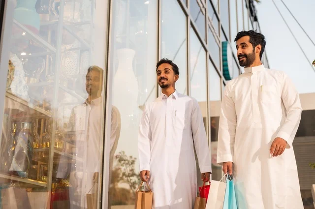 المتعة في التسوق واقتناء المستلزمات والهدايا، الاستفادة من العروضات والتخفيضات، رجلان عربيان خليجيان سعوديان يرتديان الشماغ والثوب التقليدي يحملان بيده أكياس ورقية ملونة بإيماءات السرور والسعادة