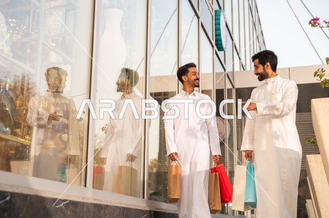 The joy of shopping and acquiring necessities and gifts, taking advantage of offers and discounts, two Arab Gulf men from Saudi Arabia wearing the traditional headscarf and thobe, holding colorful paper bags in their hands with gestures of joy and happiness.