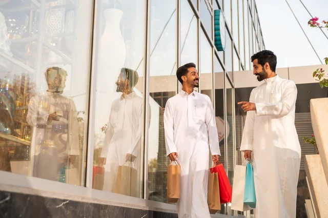 The joy of shopping and acquiring necessities and gifts, taking advantage of offers and discounts, two Arab Gulf men from Saudi Arabia wearing the traditional headscarf and thobe, holding colorful paper bags in their hands with gestures of joy and happiness.