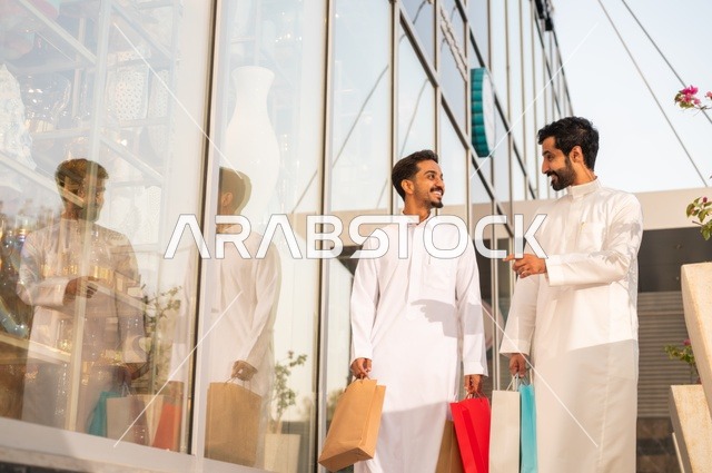 The joy of shopping and acquiring necessities and gifts, taking advantage of offers and discounts, two Arab Gulf men from Saudi Arabia wearing the traditional headscarf and thobe, holding colorful paper bags in their hands with gestures of joy and happiness.
