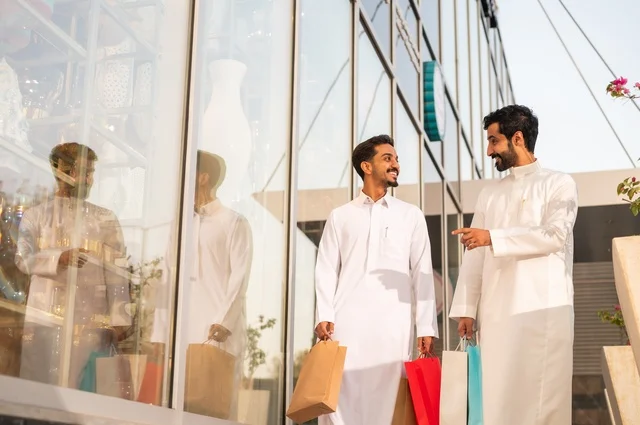 The joy of shopping and acquiring necessities and gifts, taking advantage of offers and discounts, two Arab Gulf men from Saudi Arabia wearing the traditional headscarf and thobe, holding colorful paper bags in their hands with gestures of joy and happiness.