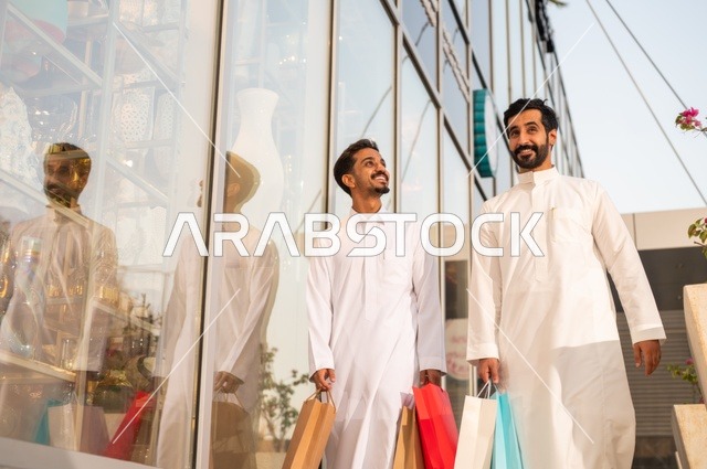 المتعة في التسوق واقتناء المستلزمات والهدايا، الاستفادة من العروضات والتخفيضات، رجلان عربيان خليجيان سعوديان يرتديان الشماغ والثوب التقليدي يحملان بيده أكياس ورقية ملونة بإيماءات السرور والسعادة