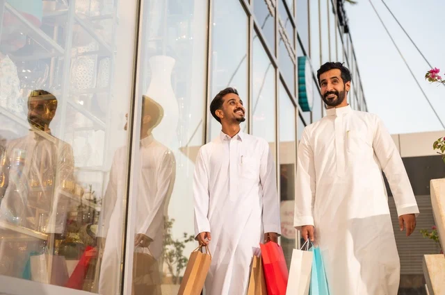 المتعة في التسوق واقتناء المستلزمات والهدايا، الاستفادة من العروضات والتخفيضات، رجلان عربيان خليجيان سعوديان يرتديان الشماغ والثوب التقليدي يحملان بيده أكياس ورقية ملونة بإيماءات السرور والسعادة