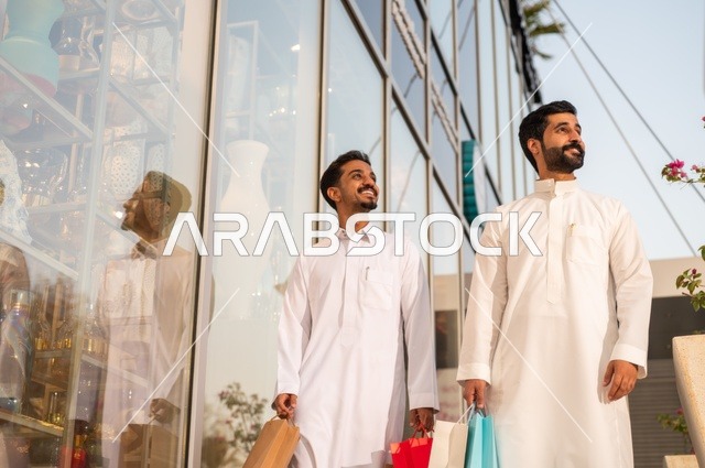 المتعة في التسوق واقتناء المستلزمات والهدايا، الاستفادة من العروضات والتخفيضات، رجلان عربيان خليجيان سعوديان يرتديان الشماغ والثوب التقليدي يحملان بيده أكياس ورقية ملونة بإيماءات السرور والسعادة