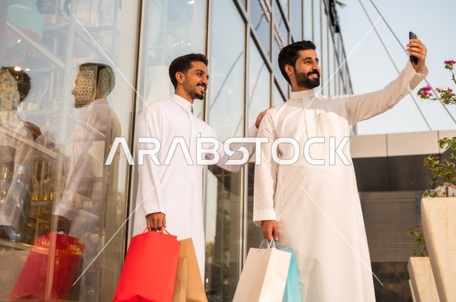 التقاط صور سيلفي شخصية باستخدام الهاتف المحمول، المتعة في التسوق واقتناء المستلزمات والهدايا، الاستفادة من العروضات والتخفيضات، رجلان عربيان خليجيان سعوديان يرتديان الشماغ والثوب التقليدي يحملان بيده أكياس ورقية ملونة بإيماءات السرور والسعادة