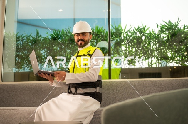 الاطلاع على تصاميم هندسية جديدة على جهاز الحاسوب المحمول، مهندس معماري عربي سعودي خليجي يرتدي اللباس الخاص بالعمل يستخدم اللابتوب، الاشراف الالكتروني على تطبيق المشاريع و الخطط الهندسيه المعمارية في البناء، دمج العمل مع التقنية و التكنولوجيا