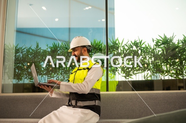 الاطلاع على تصاميم هندسية جديدة على جهاز الحاسوب المحمول، مهندس معماري عربي سعودي خليجي يرتدي اللباس الخاص بالعمل يستخدم اللابتوب، الاشراف الالكتروني على تطبيق المشاريع و الخطط الهندسيه المعمارية في البناء، دمج العمل مع التقنية و التكنولوجيا