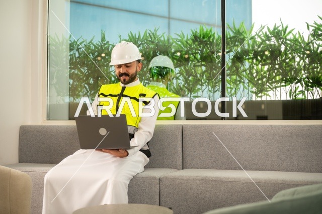 الاطلاع على تصاميم هندسية جديدة على جهاز الحاسوب المحمول، مهندس معماري عربي سعودي خليجي يرتدي اللباس الخاص بالعمل يستخدم اللابتوب، الاشراف الالكتروني على تطبيق المشاريع و الخطط الهندسيه المعمارية في البناء، دمج العمل مع التقنية و التكنولوجيا