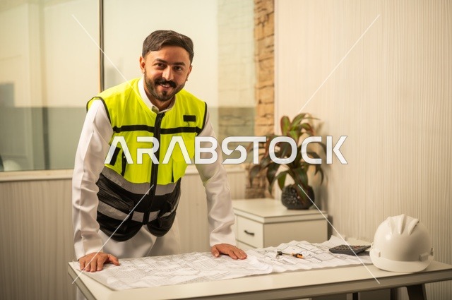 تدقيق ومراجعة الخطط الهندسية، دراسة أساسيات المشروع، إكمال رسم مخطوطات البناء، الإعمار بسواعد أبناء الوطن، مفهوم الهندسة والإنشاءات،  مهندس عربي خليجي سعودي يرتدي زي العمل يمسك بيده مخطط التطور العمراني للمباني