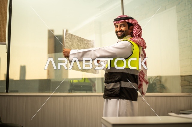 تدقيق ومراجعة الخطط الهندسية، دراسة أساسيات المشروع، إكمال رسم مخطوطات البناء، الإعمار بسواعد أبناء الوطن، مفهوم الهندسة والإنشاءات،  مهندس عربي خليجي سعودي يرتدي زي العمل يمسك بيده مخطط التطور العمراني للمباني