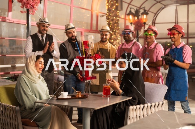 قامة حفلات اعياد الميلاد في الكافيهات والمطاعم الفاخرة في المملكة، التزيين  للاحتفال بالمناسبات السعيدة، إمشاركة طاقم الخدمة الاحتفال، قضاء وقت ممتع مع الأصدقاء، إيماءات السعادة والمفاجأة، امرأة عربية خليجية سعودية محجبة ترتدي العباءة تقوم بالاحتفال بعيد ميلاد صديقتها