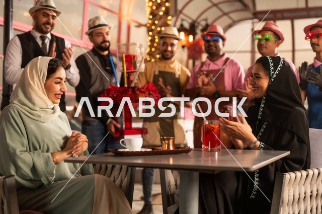 قامة حفلات اعياد الميلاد في الكافيهات والمطاعم الفاخرة في المملكة، التزيين  للاحتفال بالمناسبات السعيدة، إمشاركة طاقم الخدمة الاحتفال، قضاء وقت ممتع مع الأصدقاء، إيماءات السعادة والمفاجأة، امرأة عربية خليجية سعودية محجبة ترتدي العباءة تقوم بالاحتفال بعيد ميلاد صديقتها