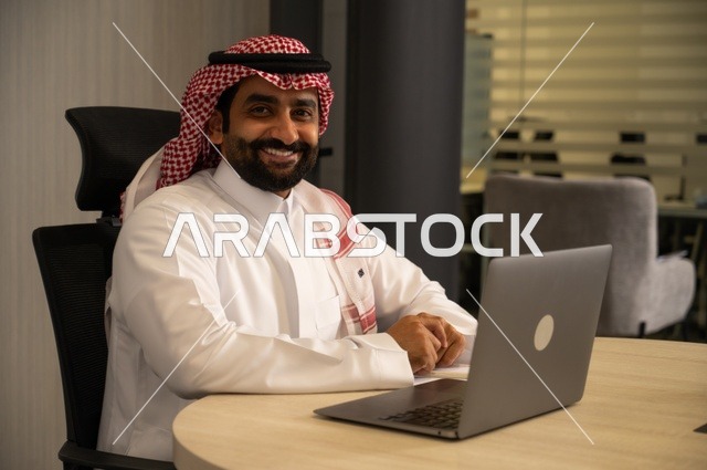 مهن وأعمال مكتبية، توفير بيئة عمل مريحة ومناسبة، استخدام جهاز الحاسوب المحمول لإنجاز المهام باحترافية، رجل عربي خليجي سعودي يرتدي الثوب والشماغ التقليدي يجلس في المكتب يقوم بالعمل عبر اللابتوب، التوظيف في المكاتب والشركات السعودية