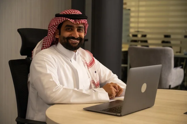 مهن وأعمال مكتبية، توفير بيئة عمل مريحة ومناسبة، استخدام جهاز الحاسوب المحمول لإنجاز المهام باحترافية، رجل عربي خليجي سعودي يرتدي الثوب والشماغ التقليدي يجلس في المكتب يقوم بالعمل عبر اللابتوب، التوظيف في المكاتب والشركات السعودية